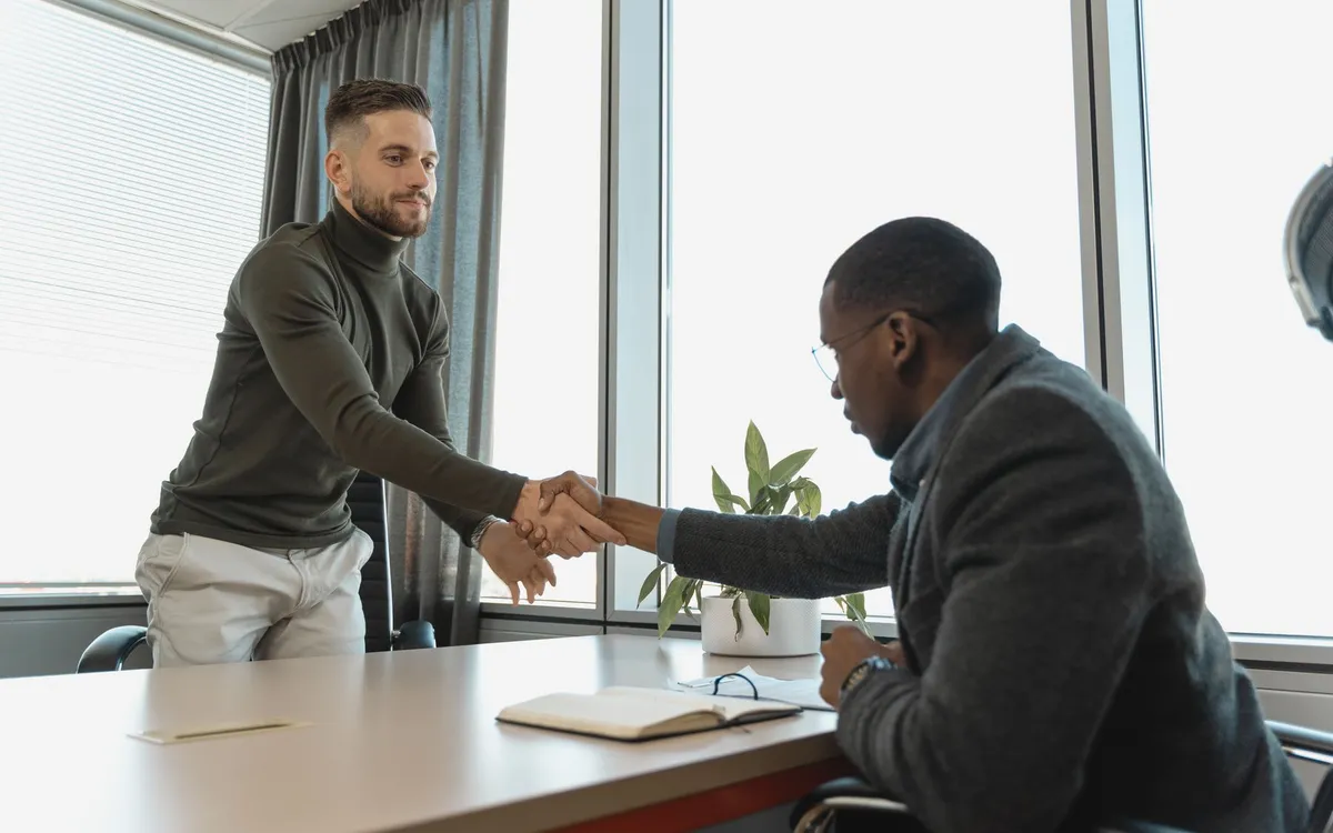 Dois homens se cumprimentando com um aperto de mão numa mesa de escritório, para o texto em inglês sobre duas propostas de emprego com comparativo