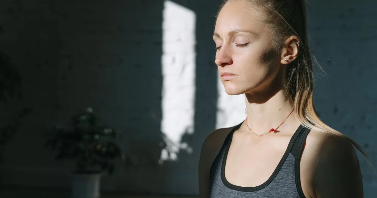 Mulher de olhos fechados respirando fundo, com calma, sob a luz de uma janela — o suspiro fisiológico na prática