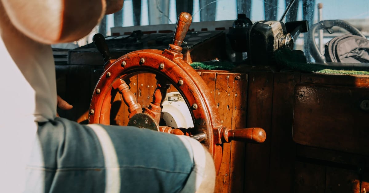 Homem ao leme de um barco, com a roda de madeira em primeiro plano, metáfora de assumir a direção da própria vida.