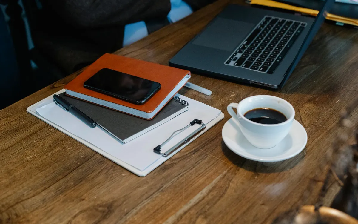 Mesa de trabalho com um laptop, uma xícara de café, dois cadernos e um celular: there is a laptop, there are two notebooks
