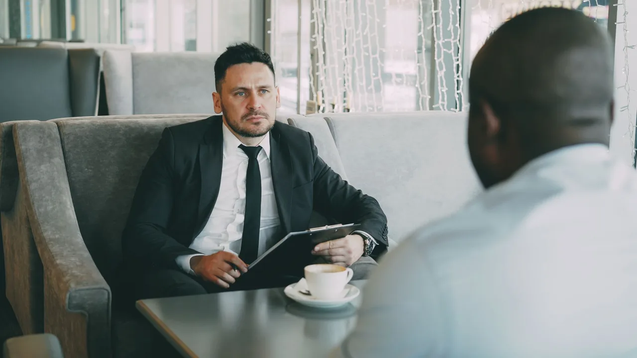 Dois executivos em conversa séria frente a frente numa mesa, um deles com uma pasta na mão