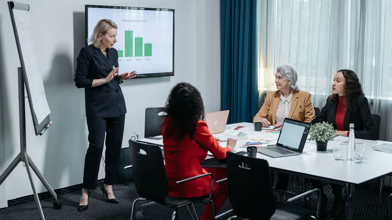 Mulher apresentando um gráfico numa tela para três colegas sentados à mesa de reunião