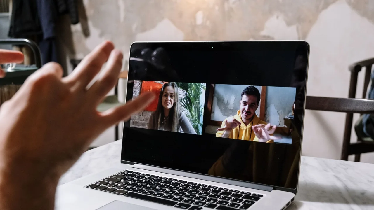 Mão gesticulando diante de um notebook com duas pessoas numa videochamada