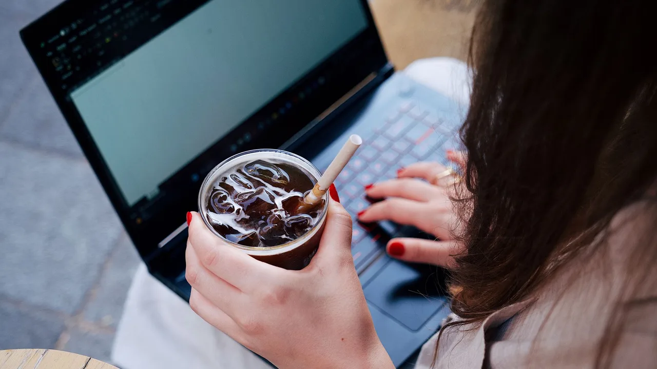 Mulher trabalhando no laptop num café, segurando um copo de café gelado