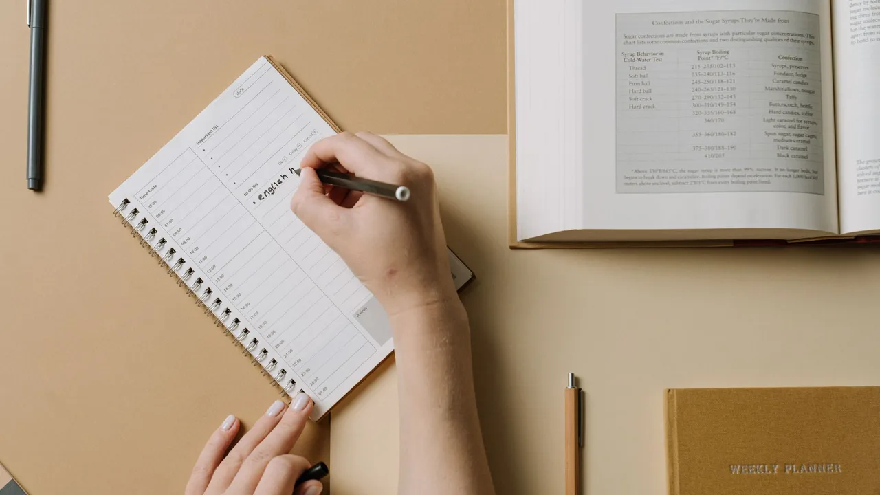 Mãos escrevendo numa agenda semanal sobre a mesa, ao lado de um livro aberto